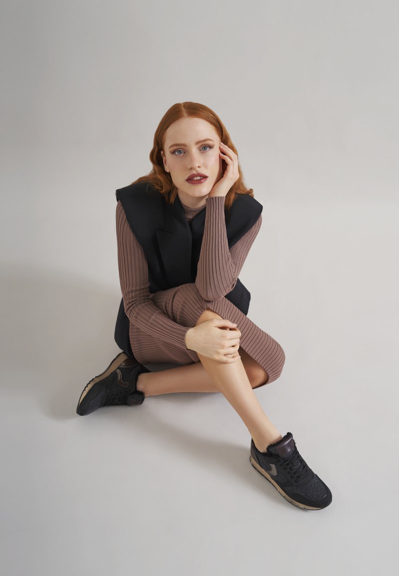 Schwarze sportliche Sneakers aus strukturiertem Material, kombiniert mit einem leicht braunen, langärmeligen Kleid mit Rippstruktur. Das Model sitzt lässig vor einem neutralen Hintergrund.