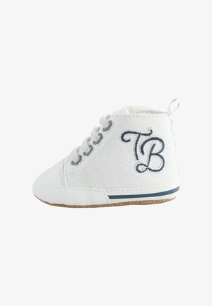Zapato de tela blanca para bebé con cordones blancos, letras bordadas "TB" en el lateral y suela de goma con raya azul marino.
