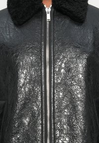 Casaco de couro preto com acabamento texturizado, fecho de zip e colarinho de shearling preto. Duas bolsos laterais são visíveis.