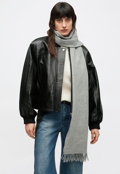 Μαύρο δερμάτινο μπουφάν τύπου bomber με μπροστινό φερμουάρ, ριμπ μανσέτες, και ανοιχτό γκρι φουλάρι με κρόσσια γύρω από τον λαιμό. Ανοιχτό μπλε τζιν παντελόνι.