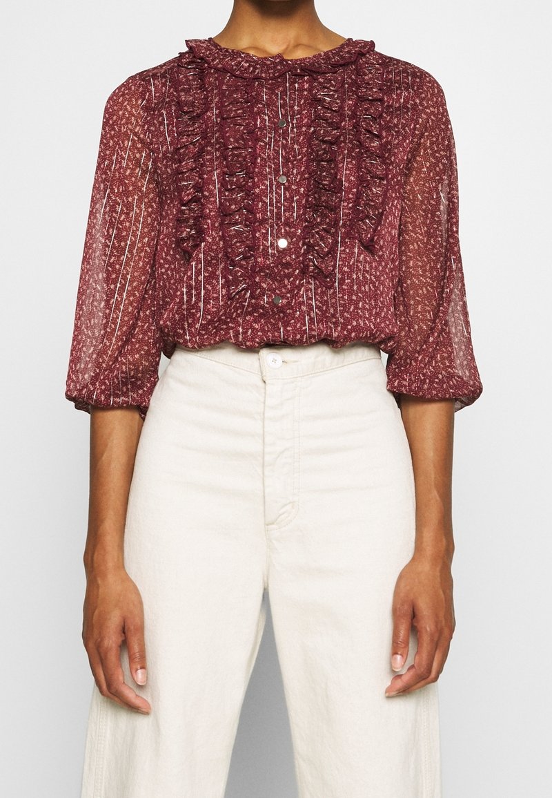 Blusa trasparente borgogna con colletto e parte anteriore arricciati, righe verticali e bottoni. Abbinata a pantaloni a vita alta di colore chiaro.