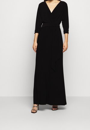 Femme portant une longue robe portefeuille noire à manches trois-quarts avec une ceinture à la taille, associée à des sandales noires à talons hauts avec des brides à la cheville.