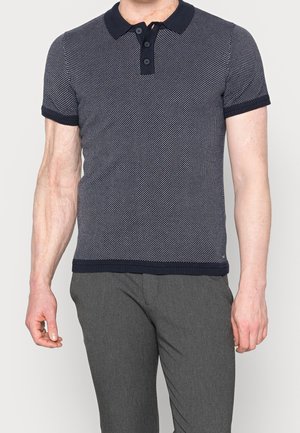 Man in een marineblauwe geweven polo met korte mouwen en een grijze broek, staand tegen een effen lichte achtergrond.