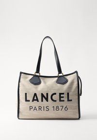 Plátěná kabelka s béžovým tělem a černými koženými detaily a uchy; výrazný černý text zní "LANCEL PARIS 1876."