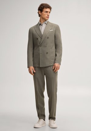 Mężczyzna ubrany w jasnoszary dwurzędowy garnitur z dopasowanymi spodniami, szarą koszulę, białą poszetkę i białe trampki, stojący na jednolitym tle.