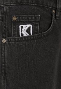 Schwarze Jeans aus Denim mit einer Fronttasche und einem weißen, gestickten Logo. Sichtbare Metallnieten und eine strukturierte Stoffoberfläche.