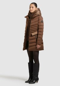 Brauner Steppmantel mit hohem Kragen, gestepptem Design und Reißverschluss vorne. Verfügt über eine verstellbare Kapuze und seitliche Taschen. Getragen mit schwarzen knielangen Stiefeln.