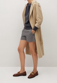 Béžový trenčkot s ohrnutými rukávy, námořnický svetr, šedé šortky a hnědé mokasíny s zlatým řetízkovým detailem. Hladké textury, ležérní vzhled.
