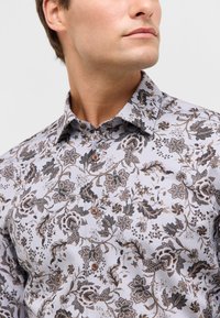 Herenoverhemd in lichtgrijs met een donker bloemenpatroon. Kenmerkt zich door een spread collar, bruine knopen en een gestructureerde stof.