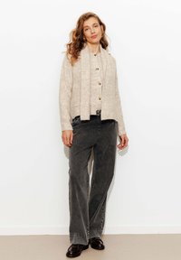 Femme aux cheveux ondulés portant un cardigan beige et une écharpe, un jean large gris foncé et des mocassins foncés, se tenant devant un fond blanc.