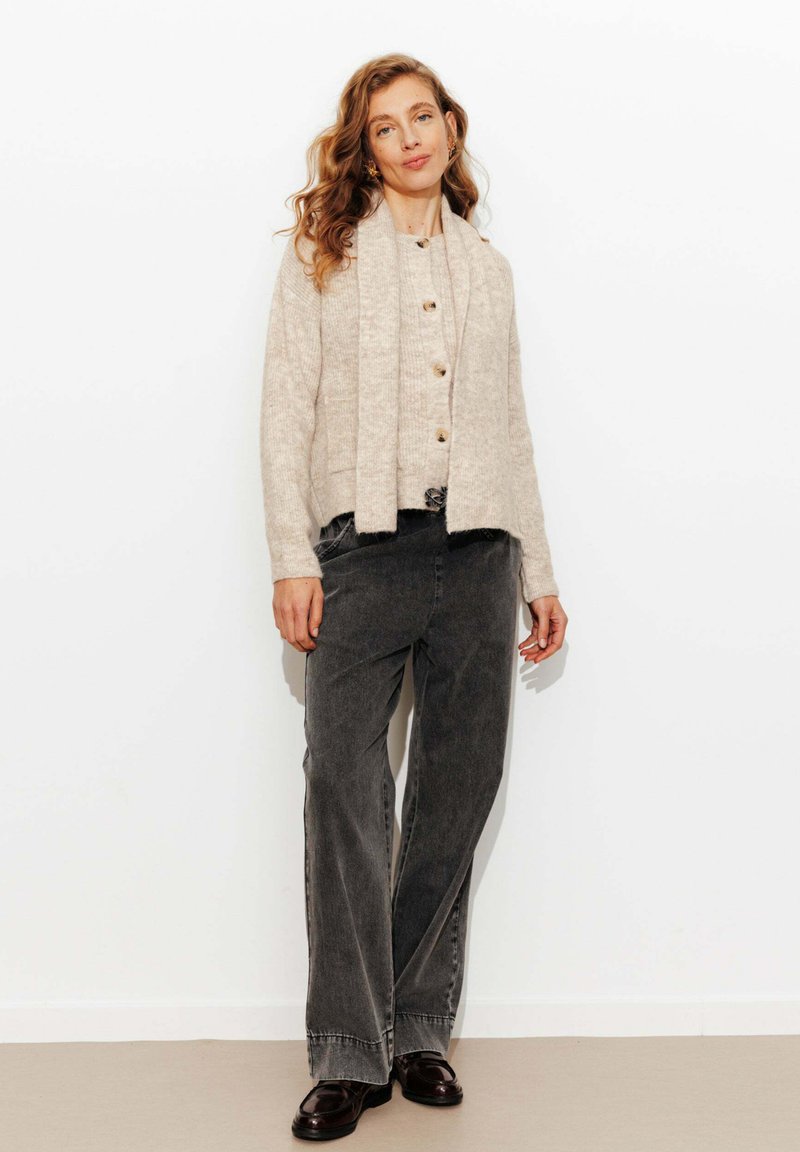 Femme aux cheveux ondulés portant un cardigan beige et une écharpe, un jean large gris foncé et des mocassins foncés, se tenant devant un fond blanc.