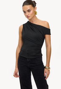 Haut noir à une épaule en tissu extensible avec des détails drapés et une silhouette ajustée, associé à un jean noir taille haute.
