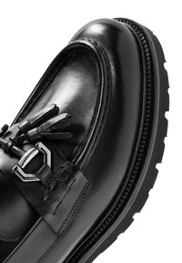 Zapato mocasín de cuero negro con punta redondeada, borlas decorativas y suela de goma gruesa en contraste con costuras visibles.