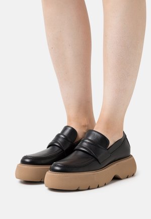 Mocassins en cuir noir avec une épaisse semelle en caoutchouc tan rainurée et une large sangle à l'avant. Texture lisse et design à bout arrondi.