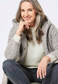 Grau- und cremefarbener Strickcardigan mit Kapuze, der ein strukturiertes Muster und große Knöpfe aufweist, getragen über einem hellcremefarbenen gerippten Pullover.