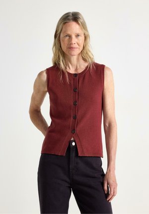 Donna con i capelli lunghi e biondi indossa una canottiera rossa con bottoni e pantaloni neri su uno sfondo chiaro e uniforme.