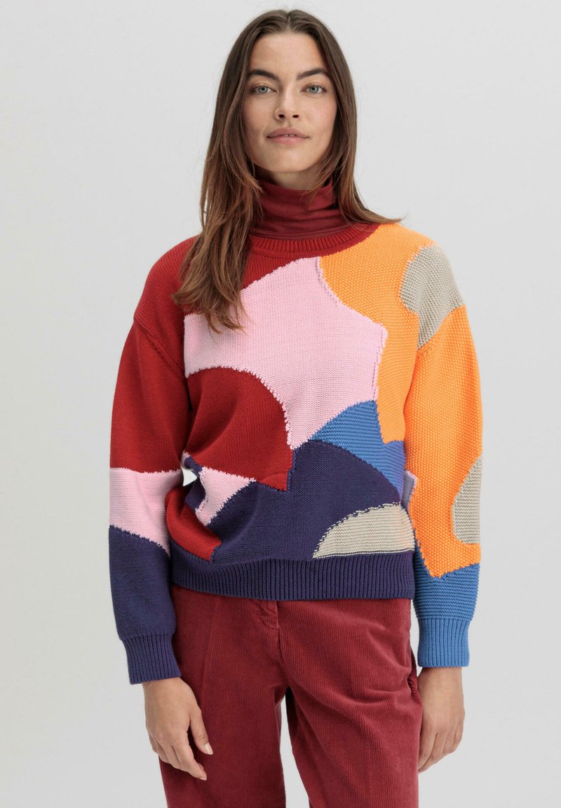 Pull en tricot coloré avec un col roulé, orné de motifs abstraits dans les teintes rouge, rose, orange, bleu et beige. Texture douce.