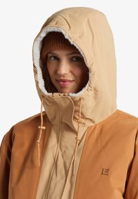 Beige och tan huva jacka med fodrad krage, dragsko och en liten logopatch på bröstet. Mjuk textur och lagerdesign.