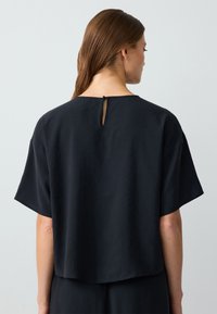 Top nero a maniche corte con una vestibilità morbida, caratterizzato da un collo rotondo e un piccolo dettaglio a occhiello sul retro. Tessuto dalla consistenza liscia.