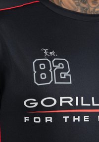 Zwarte sportshirt met rode accenten, voorzien van witte tekst "GORILLA FOR THE" en "Est. 82" in grote grijze letters. Gladde stoftextuur.