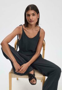 Zwarte, mouwloze jumpsuit met een ronde hals. Heeft verstelbare banden en losse broekspijpen. Draagt met zwarte, bandjes sandalen.