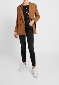 Brauner übergroßer Blazer über einem schwarzen gemusterten Top, kombiniert mit schwarzen Skinny-Jeans und weißen Sneakers. Texturierte Materialien mit glatter Oberfläche.