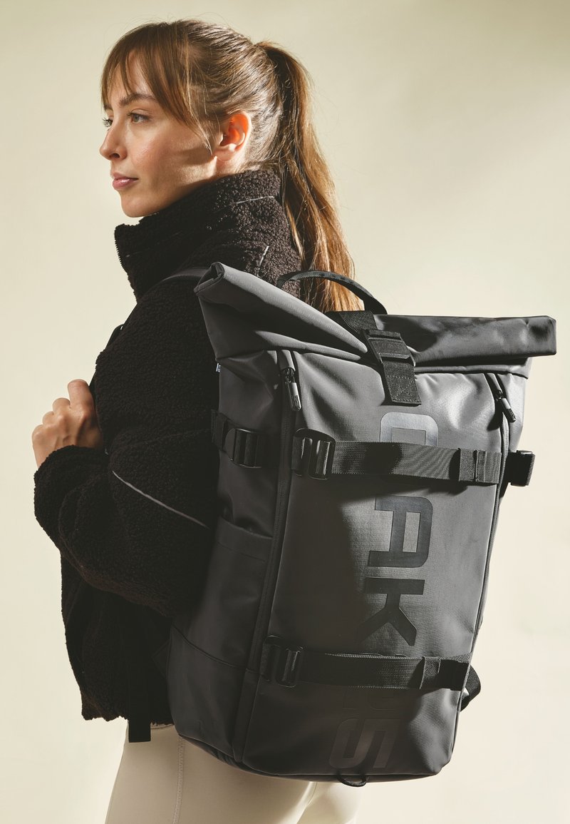 Mochila negra tipo roll-top con correas y hebillas ajustables. Fabricada con material duradero que presenta un gran logo. Textura suave, diseño funcional.