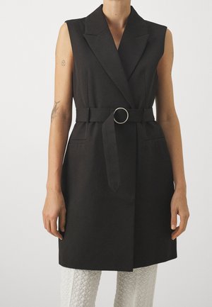 Veste noire sans manches ceinturée avec de larges revers portée sur un pantalon blanc texturé, montrant des bras avec de petits tatouages.