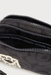 Μαύρο υφασμάτινο τσαντάκι με ανθικό μοτίβο, δύο ανοιχτές θήκες με φερμουάρ και την επωνυμία «marimekko» στην εσωτερική επένδυση και μπροστά.