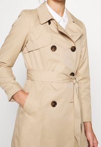 Trench beige avec un design croisé boutonné, de grands boutons foncés, une taille ceinturée et des poches latérales. Texture lisse et col pointu.