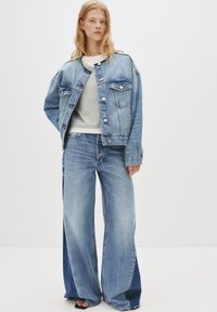 Jeans jakna svetlo modre barve s sproščenim krojem, ki ima dva prsna žepa in srebrne gumbe. V kombinaciji z širokimi svetlo modrimi kavbojkami.