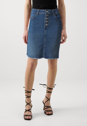 Woman wearing a blue denim button-front skirt with frayed hem and black strappy high-heeled sandals, standing on a plain background.