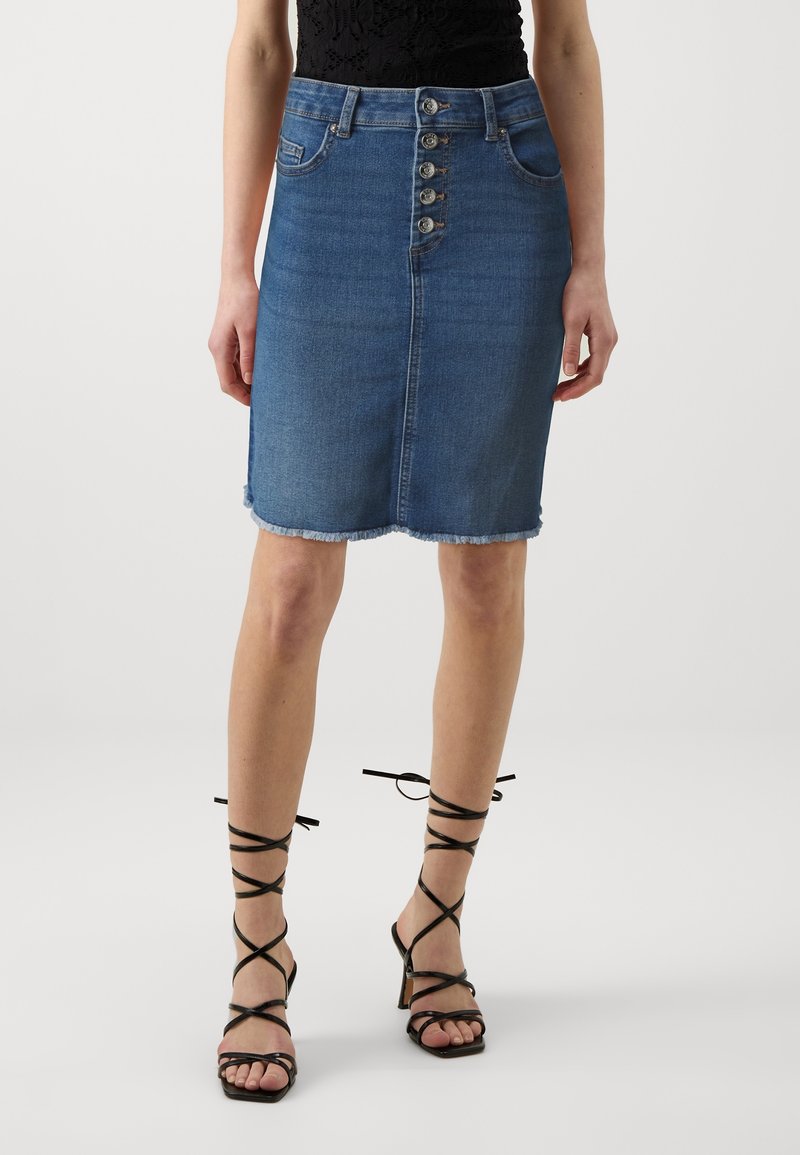 Femme portant une jupe en denim bleu à boutons sur le devant avec un ourlet effiloché et des sandales noires à talons hauts à lanières, debout sur un fond uni.