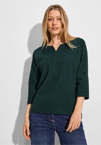 Haut texturé vert foncé avec un col en V et des manches trois quarts, associé à un jean foncé. Design simple avec un motif subtil.