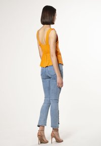 Top peplum arancione senza maniche con schiena scoperta, abbinato a jeans in denim blu chiaro, caratterizzati da un design aderente e dettagli con bottoni laterali.