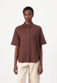 Chemise brune à manches courtes en lin avec un col, des boutons sur le devant et une coupe décontractée, assortie à un pantalon clair.