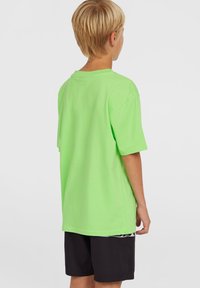 T-shirt di cotone verde neon con maniche corte e vestibilità rilassata, abbinato a pantaloni corti neri. Il tessuto appare liscio e leggero.