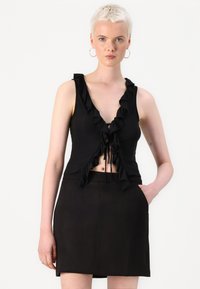 Schwarzes Crop-Top mit Rüschenausschnitt und vorderem Bindeband; kombiniert mit einem figurbetonten schwarzen Minirock mit seitlichen Taschen. Glatte Stoffstruktur.