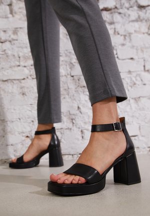 Sandalias de cuero negro de tacón alto con un tacón cuadrado grueso, punta abierta y correa en el tobillo. Textura suave, diseño minimalista y construcción robusta.