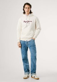 Crème hoodie met het logo "Pepe Jeans London" in bordeauxrood, met een kangoeroezak, gecombineerd met blauwe spijkerbroek en witte sneakers.