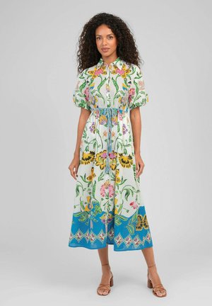 Femme debout portant une robe midi blanche avec des motifs floraux colorés, des manches bouffantes, une taille cintrée avec une ceinture tressée et des sandales marron.