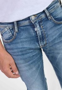Jeans in denim azzurro chiaro con un design aderente, cinque tasche e cuciture a contrasto. Il tessuto ha una texture leggermente sbiadita.
