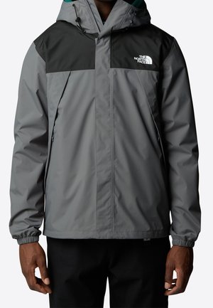 Personne portant une veste à capuche North Face grise et noire avec poignets élastiques et poches zippées, debout sur un fond blanc.