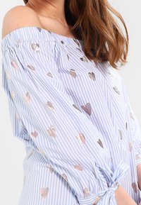 Off-shoulder blus i blå och vit randig mönster med metalliska hjärtan, med långa ärmar och knytdetalj vid midjan.