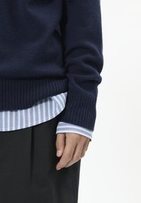 Granatowy sweter z dzianiny z prążkowanymi mankietami, nałożony na jasnoniebieską koszulę w białe paski, w połączeniu z ciemnymi spodniami z tkaniny.