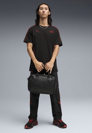 Persona joven de pie sosteniendo un maletín negro de Puma, vistiendo una camiseta negra Puma F1, pantalones negros con detalles en rojo y zapatillas negras con detalles rojos.