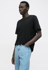 Giovane uomo dalla pelle scura che indossa una maglietta nera e jeans azzurri chiari, in piedi con una mano in tasca su uno sfondo neutro.