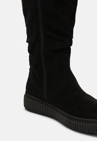 Botte haute jusqu'au genou en daim noir dotée d'une fermeture éclair latérale et d'une semelle en caoutchouc texturée avec des nervures verticales. Surface lisse et design ajusté.