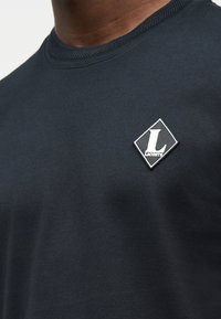 Κοντινό πλάνο σκούρου μπλε μπλουζιού με στρογγυλή λαιμόκοψη και διακοσμητική, ρομβοειδή ετικέτα με το λογότυπο Lacoste στο στήθος.