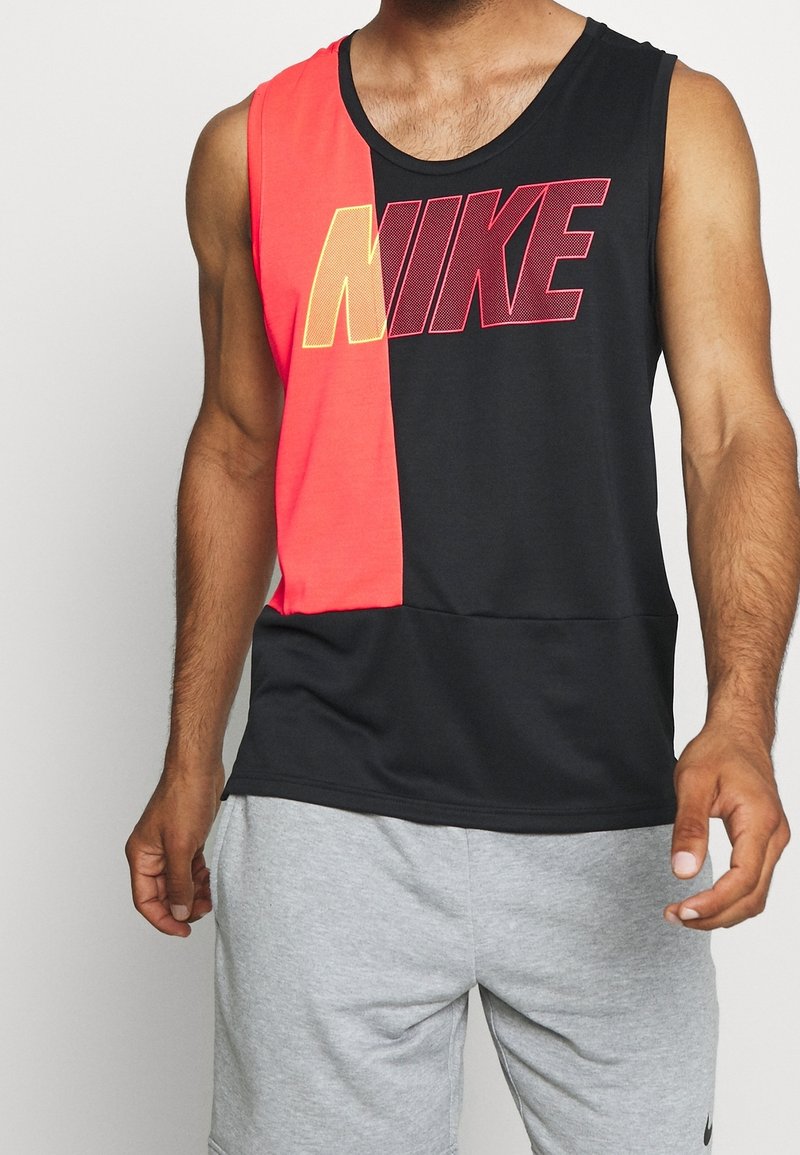Homme portant un débardeur Nike noir et rouge sans manches avec un grand logo, associé à un short gris clair.